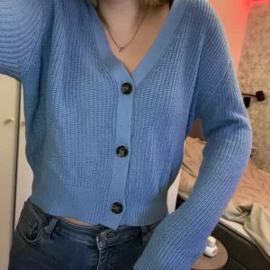 Blå stickad kofta - Säljer en superfin blå stickad kofta med stora knappar framtill. Den är perfekt för både vår och höst och har en skön, normal passform. Koftan är i ett mjukt material som känns riktigt mysigt mot huden. Perfekt att ha över en t-shirt eller linne för en avslappnad look!