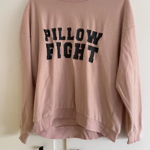 Rosa sweatshirt med texten 'Pillow Fight' - Säljer en supermjuk och bekväm rosa sweatshirt med texten 'Pillow Fight' i svart på framsidan. Tröjan har långa ärmar och en rund halsringning med ribbad kant. Perfekt för mysiga dagar hemma eller en avslappnad dag ute. Passformen är normal och materialet känns skönt mot huden.