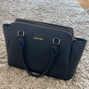 Calvin Klein väska - Svart Calvin Klein väska med gulddetaljer, använd några månader men fortfarande i bra skick. Man får även med en axelrem 