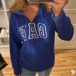 Blå hoodie från GAP - Säljer en snygg blå hoodie från GAP. Den har en dragkedja framtill och en stor logga på bröstet. Perfekt för en avslappnad stil och passar bra till jeans. Den är superbekväm och har en huva med snören. Lite slitningar 