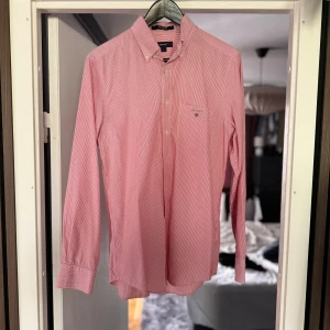 Röd- och vitrandig skjorta från GANT - Säljer en snygg röd- och vitrandig skjorta från GANT i storlek S. Skjortan har långa ärmar och en klassisk krage. Perfekt för både vardag och fest! Den är i bra skick och har en liten bröstficka med GANT-loggan broderad. Passar perfekt till jeans eller chinos för en stilren look.