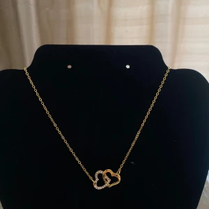 Guldhalsband med hjärtan - guldhalsband med två sammanflätade hjärtan. Det ena hjärtat är dekorerat med glittrande stenar. Perfekt för speciella tillfällen eller som en fin present. Halsbandet har en tunn kedja