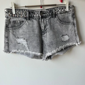 Grå jeansshorts med nitar - Säljer ett par grå jeansshorts med coola slitningar och nitar i midjan. De har en rå kant nertill och en klassisk femficksdesign. Perfekta för en avslappnad sommarlook!