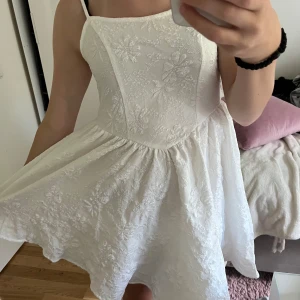 Vit blommig klänning - Säljer en superfin vit klänning med blommigt mönster. Den är ärmlös och har en söt, luftig passform som passar perfekt för sommarens alla fester. Klänningen är i nyskick och har en elegant design med en lätt volang i kjolen.Den är ganska stretsig så skulle säga att det passar en Xs-S
