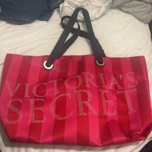Röd randig shoppingväska från Victoria's Secret - Säljer en snygg och rymlig väska från Victoria's Secret. Väskan har ett randigt mönster och bokstäver i glittriga stenar på framsidan. Den har svarta handtag. Köpt second hand. Defekter på baksidan och undersidan. Tvättar innan så ser om det går bort
