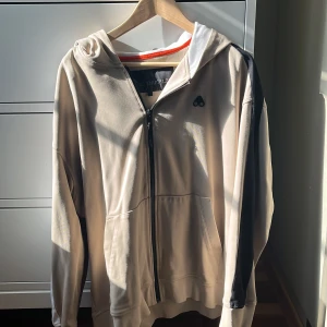 Beige hoodie från Moose Knuckles - Säljer en snygg beige hoodie från Moose Knuckles. Den har en dragkedja framtill och en cool logga på bröstet. På baksidan av huvan finns det en text som ger den en unik touch. Knappt använd. Nypris 2600kr