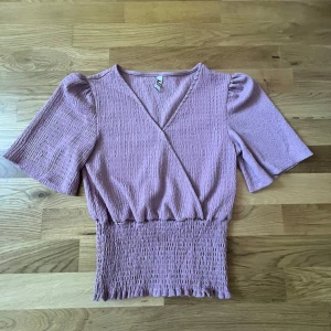 Ljuslila omlottopp - Säljer en superfin ljuslila omlottopp med korta puffärmar. Toppen har en smockad midja som ger en snygg passform och en v-ringad hals. Perfekt för både vardag och fest!