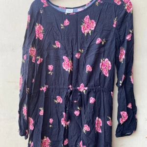 Blommig klänning från Jacqueline de Yong - Säljer en superfin blommig klänning från Jacqueline de Yong. Den är mörkblå med rosa blommor och har en avslappnad passform. Klänningen har långa ärmar och ett knytband i midjan för en snygg siluett. Perfekt för både vardag och fest!