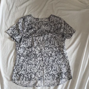 Svartvit mönstrad blus - Säljer en snygg svartvit mönstrad blus med korta ärmar. Blusen har en dragkedja i ryggen och är perfekt för både vardag och fest. Materialet känns lätt och luftigt, vilket gör den skön att bära under varmare dagar.