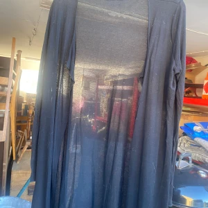 Svart lång cardigan från Vila - Säljer en svart lång cardigan från Vila. Den är gjord i ett tunt och genomskinligt material, perfekt för lager-på-lager-stil. Cardiganen har långa ärmar och en lös passform, vilket gör den superbekväm att bära över en topp eller klänning.