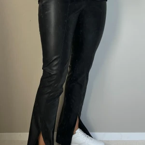 Svarta skinnbyxor med slits - Säljer ett par snygga svarta skinnbyxor med slits vid anklarna. De är tighta upptill och har en cool bootcut-stil. Perfekta för en kväll ute eller när du vill känna dig extra trendig. Passar bra med sneakers eller klackar! 