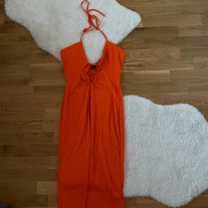 Orange långklänning från Gina Tricot - Snygg orange långklänning från Gina Tricot med knytband runt halsen och en cool detalj framtill. Perfekt för att sticka ut med sin starka färg och unika design. Klänningen har en v-ringad stil och är ärmlös, vilket gör den idealisk för varmare väder. Storlek M