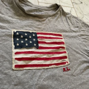 Grå t-shirt med amerikansk flagga från Polo Ralph Lauren - Säljer en grå t-shirt från Polo Ralph Lauren med en broderad amerikansk flagga på framsidan. T-shirten har korta ärmar och en rund halsringning. Den i barnstorlek L men passar en xs-s jätte bra säljer då den inte kommer till någon andvändning. Finns inga fler bilder🩷🩷 