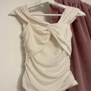 Vit topp med draperad design - Elegant vit topp med en unik draperad design och axelbandslös stil. Perfekt för speciella tillfällen med sin stilrena look.