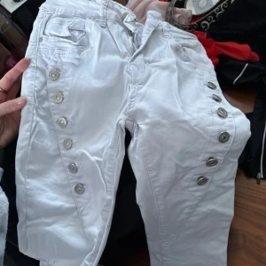 Vita byxor med knappar från Paulia - Snygga vita byxor från Paulia med dekorativa knappar längs sidorna. Byxorna har en klassisk femficksdesign och är perfekta för en stilren look. De är tillverkade i ett bekvämt material som passar bra till vardags.