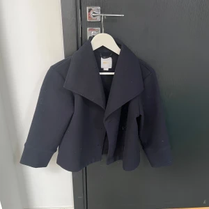 Svart kappa med bred krage - Elegant svart kappa med bred krage och knappar framtill. Perfekt för kyligare dagar med sin stilrena design. Jackan har långa ärmar och en klassisk passform.