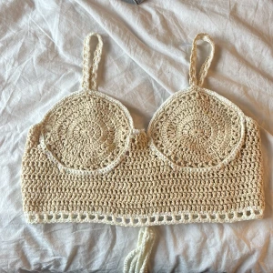 Beige virkad topp - Säljer en snygg beige virkad topp med smala axelband och snörning i ryggen. Toppen har en unik design med cirkulära mönster framtill och är perfekt för sommardagar. Då den har ett inner tyg vid brösten kan den användas utan bh💕Den är croppad och ger en bohemisk vibe💕 knappt använd därför i väldigt bra skick! Passar xs-s.