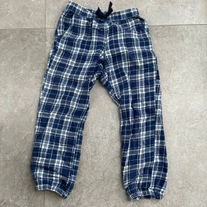 Blå och vit rutiga pyjamas byxor  - Säljer ett par bekväma blå och vit rutiga pyjamas byxor med elastisk midja och snörning. Perfekta för en avslappnad dag hemma. Byxorna har elastiska muddar vid bensluten för extra komfort.