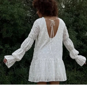 Nakd klänning - Charmig vit klänning med blommigt mönster och öppen rygg. Den har långa ärmar med knytning i ryggen och en luftig passform. Perfekt för studenten eller midsommar!