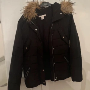 Svart dunjacka med pälsdetalj - Snygg svart dunjacka med fuskpäls på huvan. Jackan har dragkedja och knappar framtill samt två fickor med dragkedja. Perfekt för kyliga dagar.