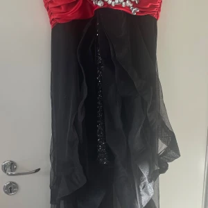 Röd och svart klänning med pärldetaljer - Elegant klänning med röd överdel och svart kjol. Den har pärldetaljer vid bysten och ett flödande tyg som ger en dramatisk look. Perfekt för speciella tillfällen.