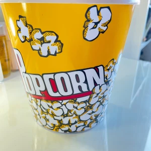 Stor gul popcornhink med tryck - Stor gul hink för popcorn med ett färgglatt tryck av poppade popcorn och texten 'Popcorn' i vitt och rött. Perfekt för filmkvällar eller biobesök. Hinken är gjord av plast och har en glansig yta.