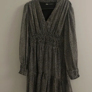 Svart och vit klänning från Zara - Snygg svart och vit klänning från Zara med v-ringning och långa ärmar. Klänningen har ett diskret mönster och smockdetaljer i midjan för en smickrande passform. Perfekt för både vardag och festliga tillfällen.