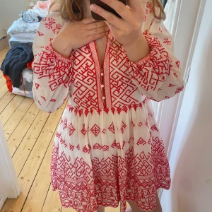 Vit och röd klänning med broderi från maje - Charmig vit klänning med röda broderade mönster. Den har långa ärmar med elastiska muddar och en v-ringad hals med knappar framtill. Perfekt för en bohemisk look. I snorkel 36