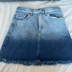 Blå jeanskjol från &Denim - Snygg blå jeanskjol från &Denim med en cool ombre-effekt. Kjolen har en klassisk femficksdesign och fransig nederkant som ger en avslappnad look. Perfekt för en trendig stil!