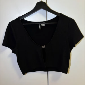 Svart ribbad kortärmad topp från H&M - Snygg svart ribbad kofta/topp från H&M med korta ärmar och en söt fjärilsdetalj framtill. Perfekt för en stilren look.