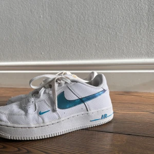 Vita Nike Air sneakers med blå detaljer - Snygga vita Nike Air sneakers med blå swoosh och detaljer. Skorna har klassisk snörning och perforeringar på ovansidan för ventilation. Perfekta för en sportig look.