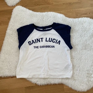 Vit och marinblå t-shirt med Saint Lucia-tryck - Säljer en cool vit och marinblå t-shirt med texten 'Saint Lucia The Caribbean' på framsidan. T-shirten har korta ärmar och en klassisk passform. Perfekt för en avslappnad stil! Liten i storlek 