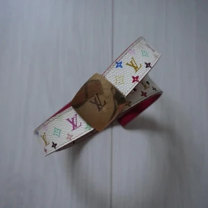 Multifärgat bälte från Louis Vuitton - Snyggt bälte från Louis Vuitton med ett färgglatt monogrammönster på vit bakgrund. Bältet har en guldig spänne med LV-logotypen och en rosa insida. Perfekt för att ge en lyxig touch till din outfit. 90x3cm
