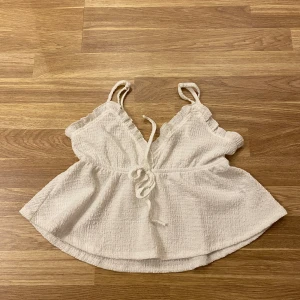 Vit ärmlös topp med knytband - Söt vit topp med smala axelband och knytband framtill. Den har en lätt textur och volangdetaljer vid axelbanden. Perfekt för varma dagar.