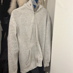 Grå hoodie med strassdetaljer från One of One - Säljer en snygg grå hoodie från One of One med glittrande strassdetaljer på framsidan. Tröjan har en dragkedja och en bekväm huva. Perfekt för en stilren och avslappnad look.