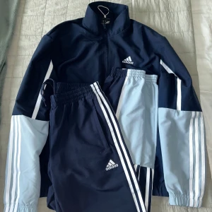 Mörkblå Tracksuit byxor och jacka från Adidas - Säljer en mörkblå tracksuit från Adidas med vita och ljusblå detaljer. Jackan har dragkedja och långa ärmar med de klassiska tre ränderna längs sidorna. Perfekt för sportiga aktiviteter eller en avslappnad stil. 🏃‍♂️