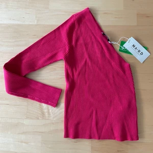 Röd ribbad topp från NA-KD M oanvänd  - Snygg röd ribbad topp från NA-KD med en unik asymmetrisk design. Perfekt för att ge din outfit en trendig touch. Toppen har långa ärmar och är tillverkad i ett mjukt material som ger en skön passform.