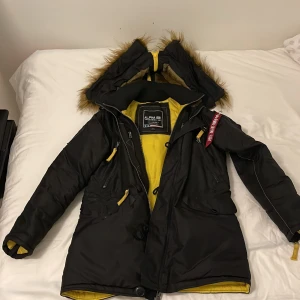 Svart parkajacka från Alpha Industries - Säljer en svart parkajacka från Alpha Industries med gul insida och pälsdetaljer på huvan. Jackan har dragkedja och knappar framtill samt flera praktiska fickor. Perfekt för kalla dagar med sin varma och skyddande design.