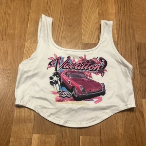 Vit croppad linnetopp med tryck - Säljer en vit croppad linnetopp med ett färgglatt tryck av en rosa bil och texten 'Vacation Enjoy'. Perfekt för en avslappnad och somrig look. Toppen har breda axelband och rund halsringning.🩷