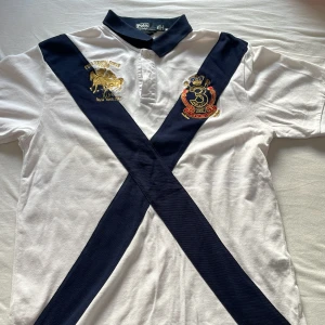 Vit pikétröja från Ralph Lauren - Säljer en stilren vit pikétröja från Ralph Lauren med mörkblå diagonala ränder och broderade emblem i guld och rött. Tröjan har en klassisk krage och knappar framtill. Perfekt för en sportig look! 100% cotton
