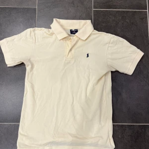 Ralph Lauren pike - Säljer en stilren beige pikétröja från Ralph Lauren med klassisk krage och korta ärmar. Tröjan har en broderad logotyp på bröstet. Perfekt för en avslappnad men ändå snygg look. Passar S/M