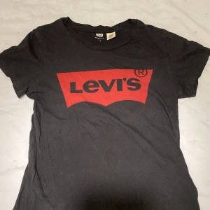 Svart t-shirt från Levi's - Säljer en klassisk svart t-shirt från Levi's med det ikoniska röda logotyptrycket på framsidan. T-shirten är kortärmad och tillverkad i mjuk bomull. Perfekt för en avslappnad stil.