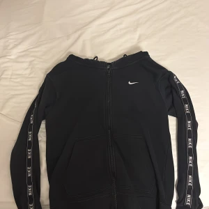 Svart hoodie från Nike - Snygg svart hoodie från Nike med dragkedja och vita logotyper längs ärmarna.