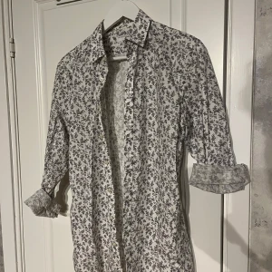 Vit blommig skjorta - Snygg vit skjorta med blommigt mönster. Perfekt för en stilren look. Skjortan har långa ärmar och knappar framtill. Passar bra till både jeans och chinos.