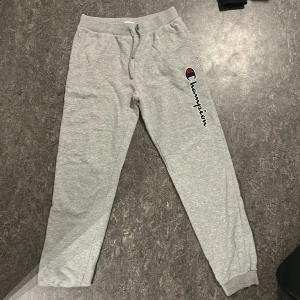 Grå mjukisbyxor från Champion - Säljer ett par grå mjukisbyxor från Champion med elastisk midja och snörning. Byxorna har en broderad Champion-logga på sidan och är perfekta för en avslappnad stil.