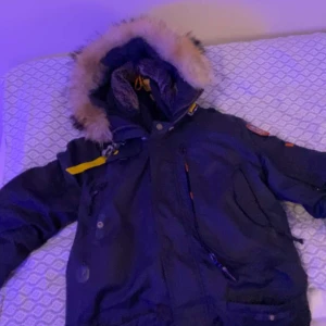 Svart parajumper  med pälsdetaljer - Säljer en svart parajacka med pälsfodrad huva och flera praktiska fickor. Jackan har en dragkedja och knappar framtill samt en dekorativ patch på ärmen. Perfekt för kalla dagar. Säljer då den ej kommer till användning 