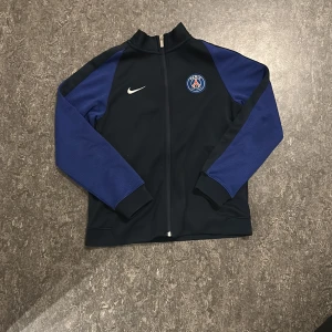 Svart och blå jacka från Nike - Snygg svart och blå jacka från Nike med PSG-logga på bröstet. Jackan har en dragkedja framtill och långa ärmar. Perfekt för fotbollsfans som vill visa sitt stöd för Paris Saint-Germain.