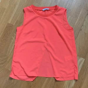 Korallfärgad ärmlös topp från J.Lindeberg - Snygg korallfärgad ärmlös topp från J.Lindeberg. Perfekt för varma dagar med sin lätta och luftiga design. Toppen har omlottdetalj bak och rund halsringning. Använd två gånger.