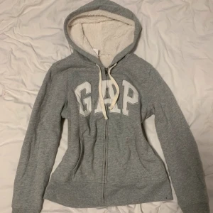 Grå hoodie från GAP - Mysig grå hoodie från GAP med dragkedja och vit text på bröstet. Den har en fodrad huva och snören för justering. Perfekt för kyliga dagar.