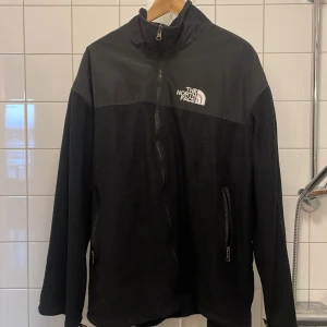 Svart fleecejacka från The North Face - Säljer en svart fleecejacka från The North Face med dragkedja och hög krage. Jackan har The North Face-logga på bröstet och är tillverkad i Gore-Tex-material för extra skydd mot väder och vind.